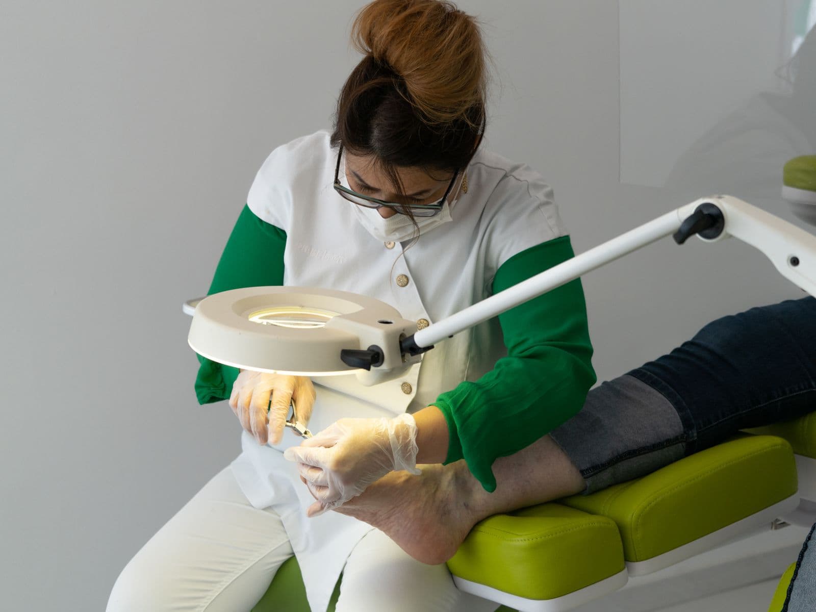 A foot-care nurse in gloves and mask treating a patient's foot under a clinic lamp.